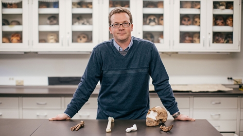 Jeremy DeSilva standing behind a table with 3D printed versions of the discovered homo naledi fossils